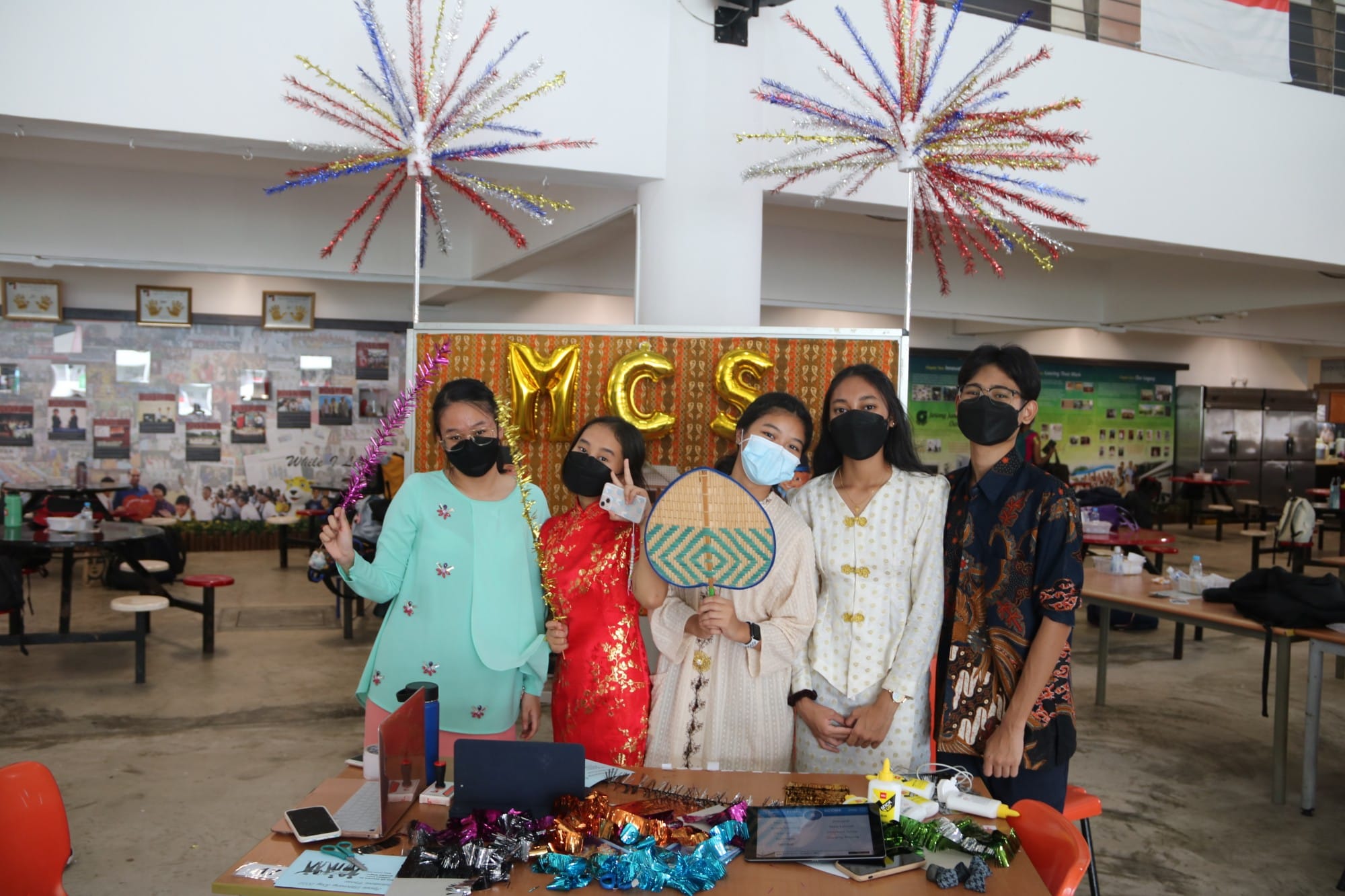 Five masked students in traditional outfits pose at a decorated booth with gold MCS balloons and colorful starburst decorations.